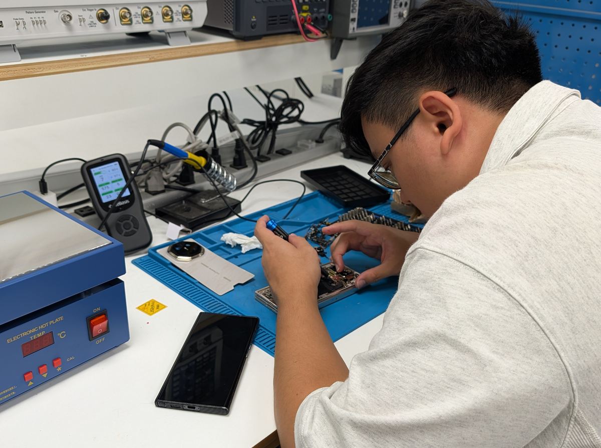 Howie at the electronics bench unscrewing a phone's frame next to a hot plate and an oscilloscope