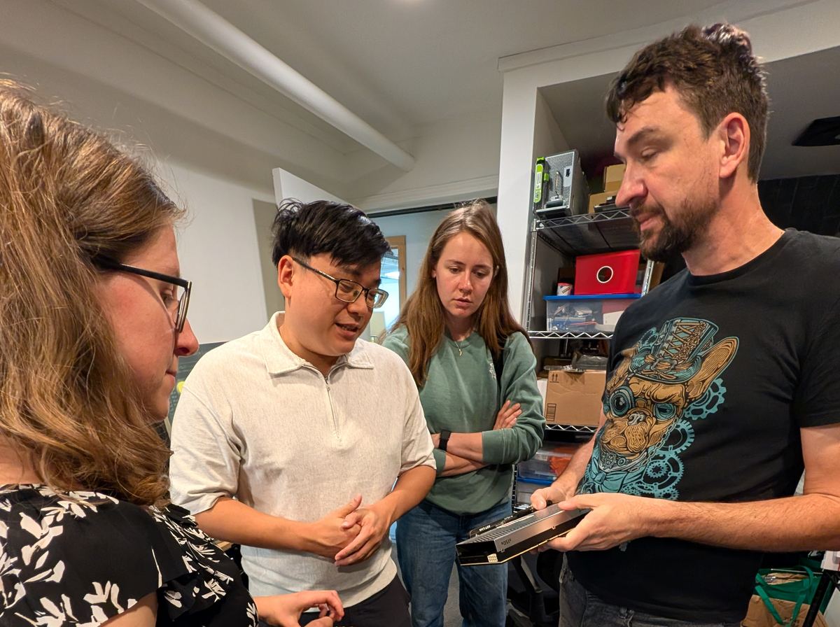 Roman holds a card while Tanya, Howie and Laura look on, beside the lab's shelves of hardware